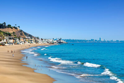 Malibu beach with people walking along the Carbon Beach shoreline, oceanfront homes in the background, and blue coastal water under clear skies — showcasing the Malibu lifestyle and luxury coastal living.