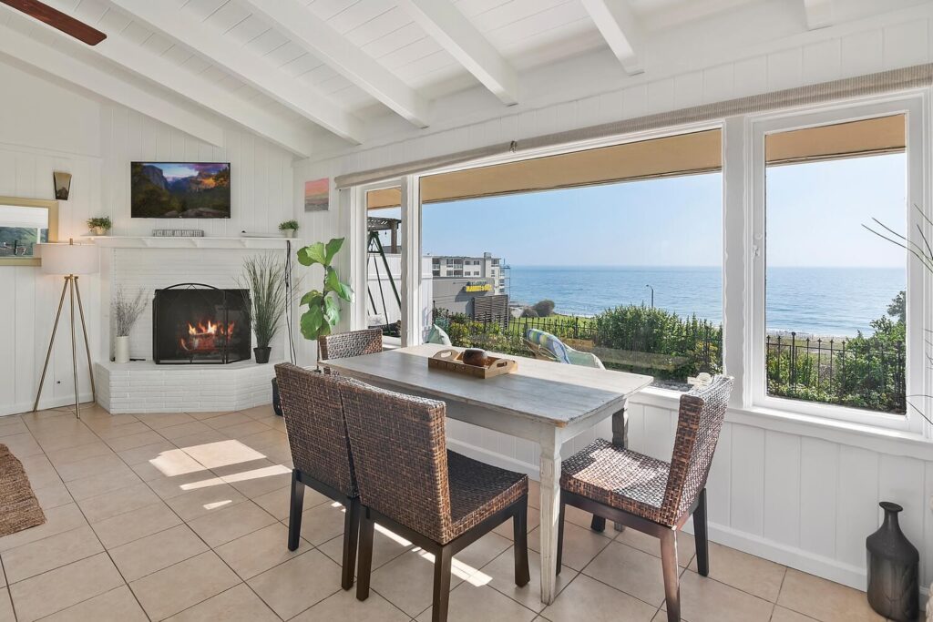 Interior of the Malibu Airbnb rental with ocean view from the dining area, white beam ceilings, fireplace, and large windows overlooking Carbon Beach.