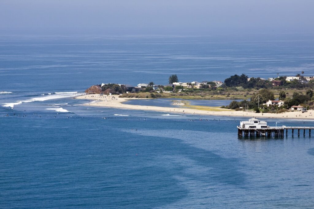 Aerial view of Malibu Pier and Carbon Beach with surfers, shoreline, and blue water on a clear day.