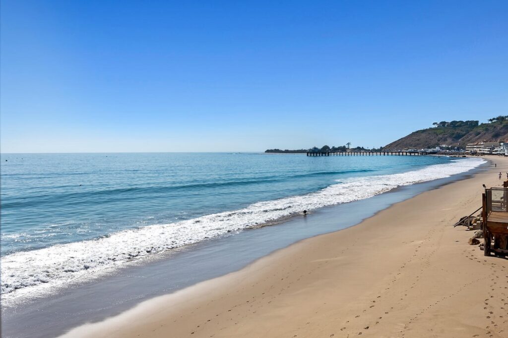 Wide sandy shoreline on Carbon Beach with calm waves, blue water, and the Malibu Pier in the distance
