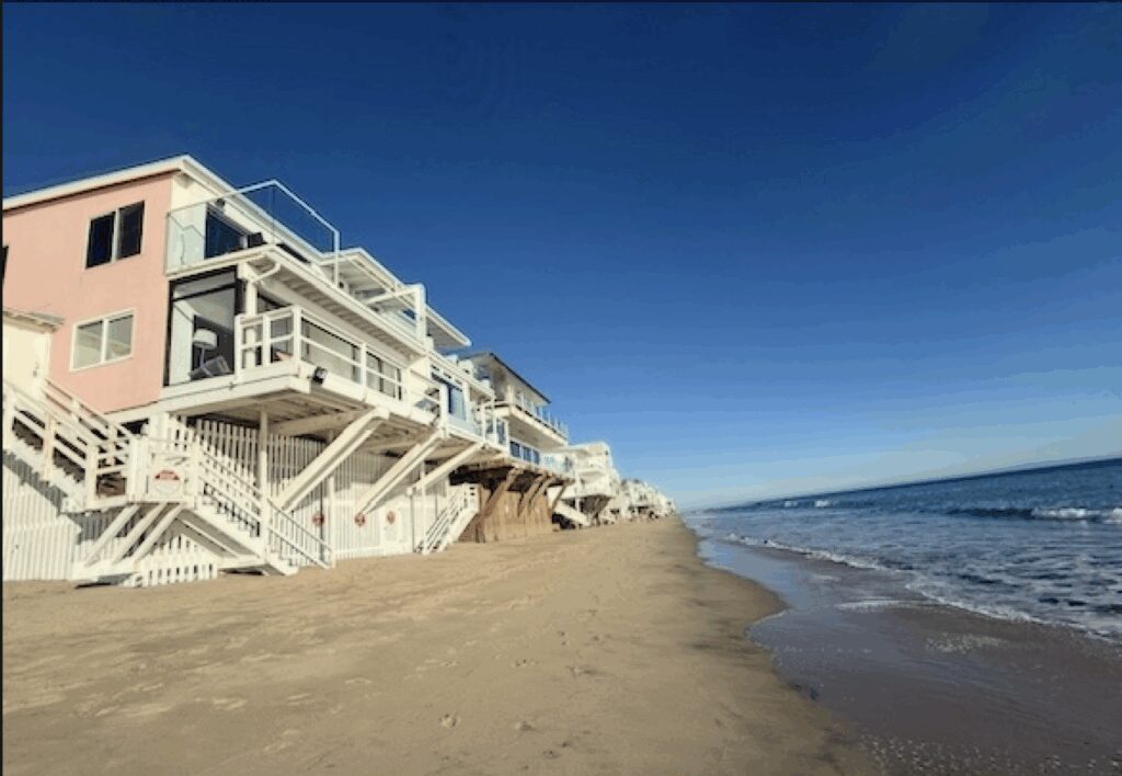 Stilted beachfront homes along Malibu Road during low tide on a clear day, showing sandy shoreline and calm waves.