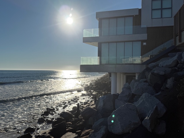 Modern oceanfront home on Malibu Road built above the rocky shoreline, captured in late-afternoon light with sun reflecting on the Pacific.
