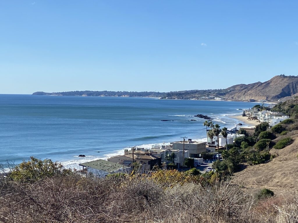 Hillside overlook view of Malibu Road’s beachfront homes and coastline on a clear day.