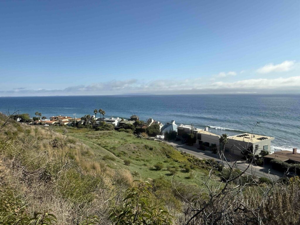 Wide hillside view overlooking Malibu Road’s beachfront homes and the Pacific Ocean.