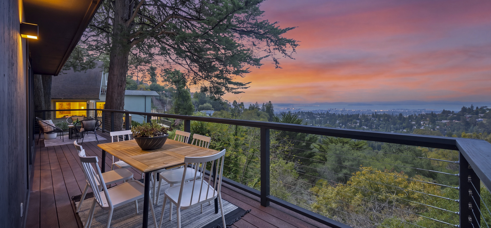 Outdoor dining deck with lounge chairs beneath a tall tree, overlooking the Oakland hills and city lights at dusk