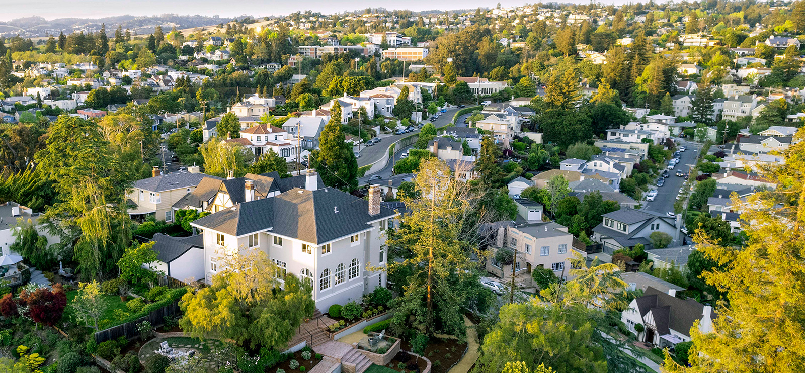 Aerial view of a white stucco home with arched windows and terraced gardens on a wooded hillside in Piedmont, California