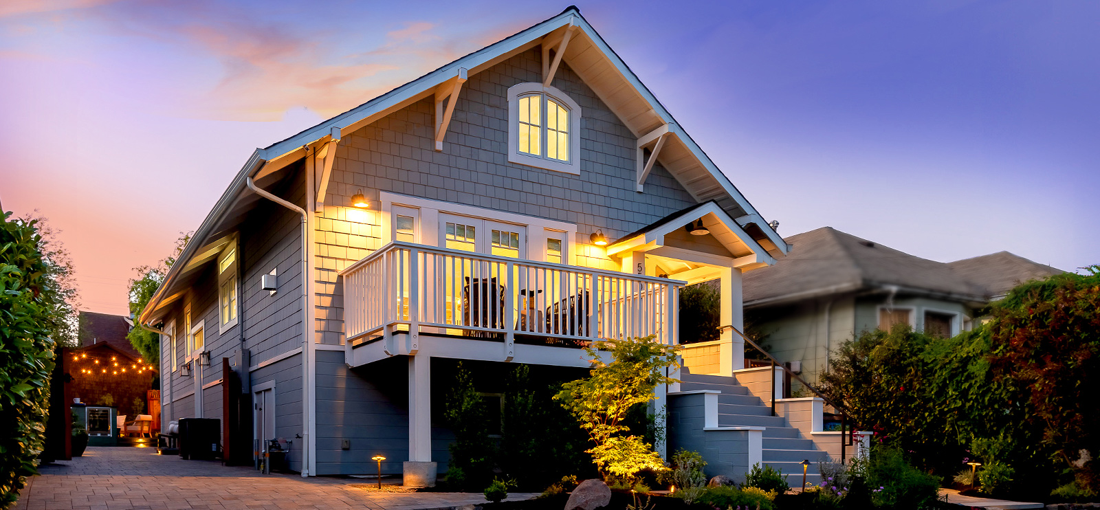 Gray shingled Craftsman home at twilight with a lit balcony, covered front porch and landscaped garden in Rockridge, Oakland
