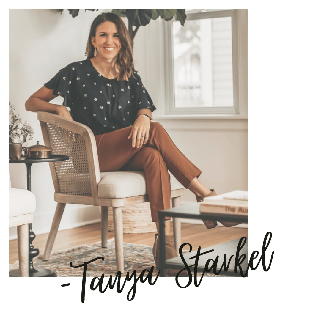 A woman sits in a chair, smiling, wearing a black top with patterns and brown pants. A coffee table and plants are nearby.