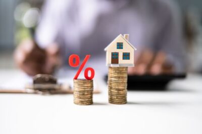 Stacks of coins with a red percent symbol and a miniature house representing interest rate changes and mortgage costs.