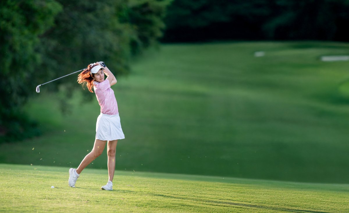 A woman in a pink shirt and white skirt is swinging a golf club